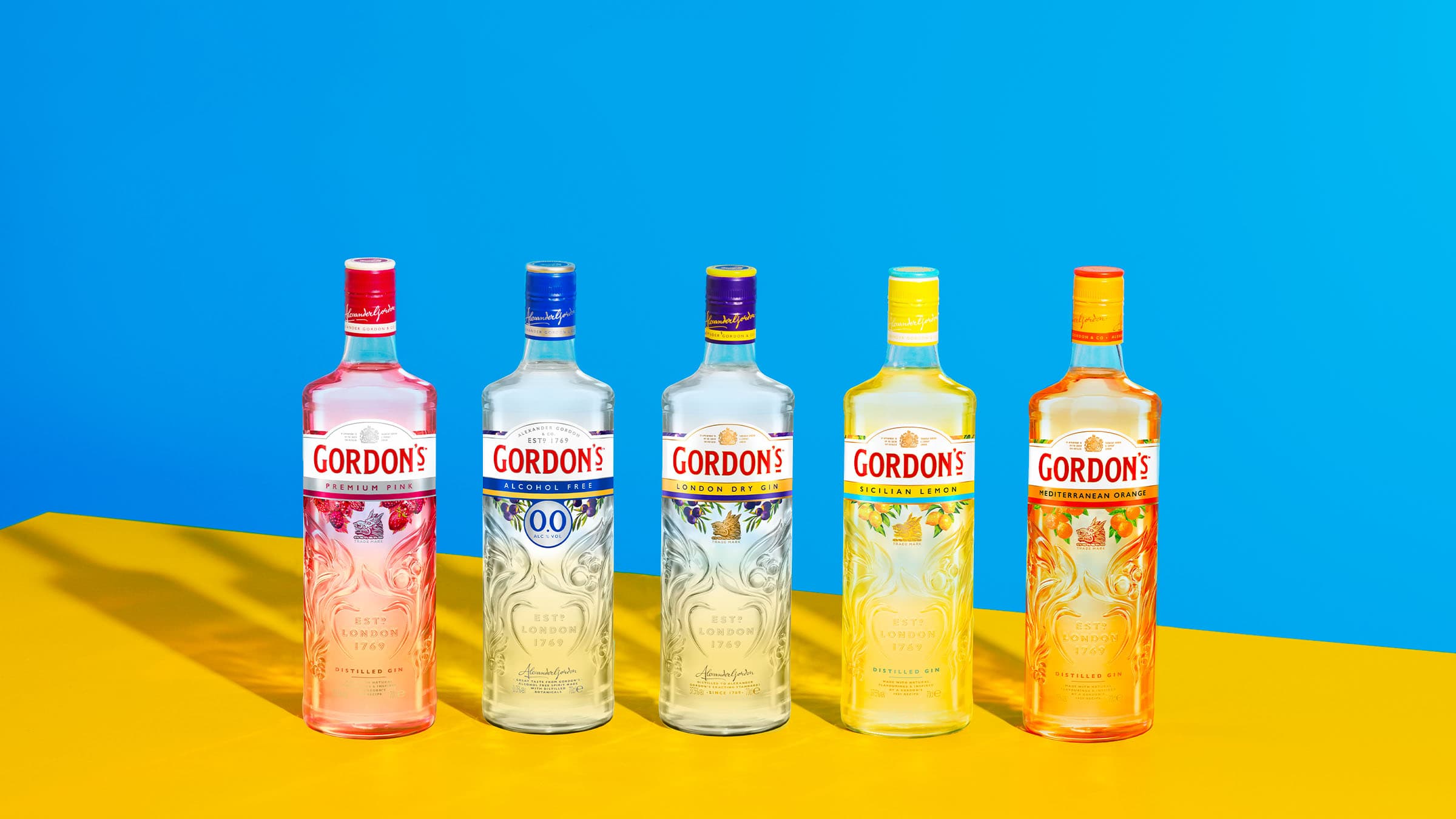 Cinco garrafas de diversas variações de gin Gordon's em um fundo azul e amarelo brilhante