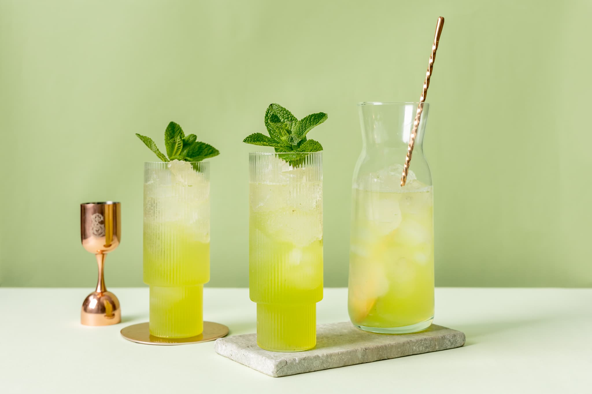 Image of two green cocktails on a table with a glass jug of the same cocktail