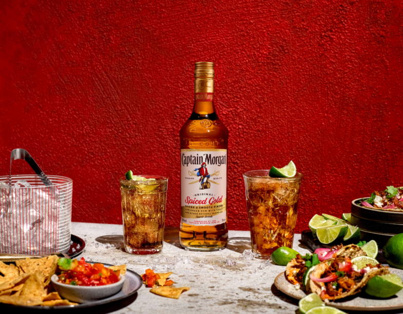 Bottle of Captain Morgan Spiced Gold rum on table beside plates of food against red background 