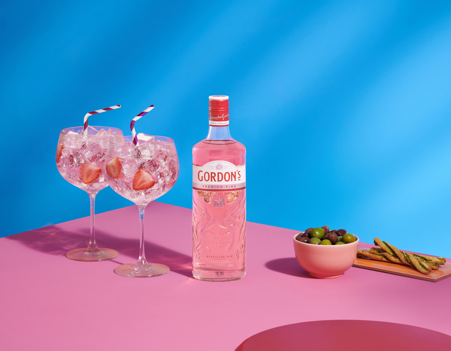 Bottle of Gordon’s Premium Pink gin beside two G&T glasses against pink and blue background