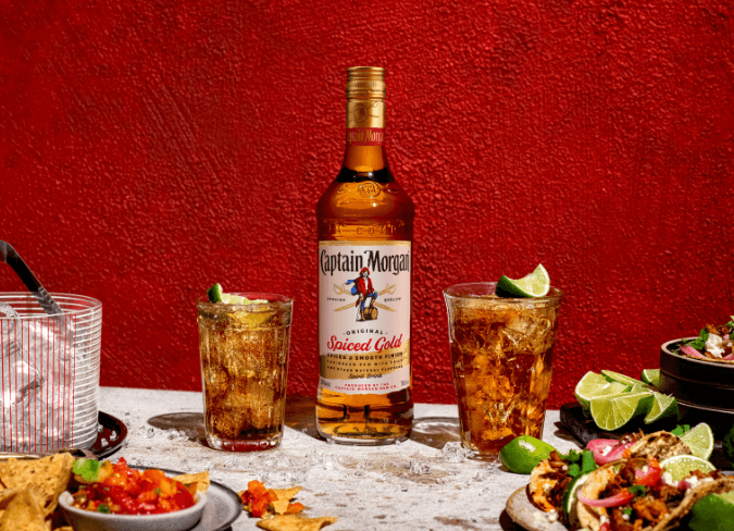 Botella de ron Captain Morgan Spiced Gold encima de una mesa junto a unos platos de comida, sobre un fondo rojo 