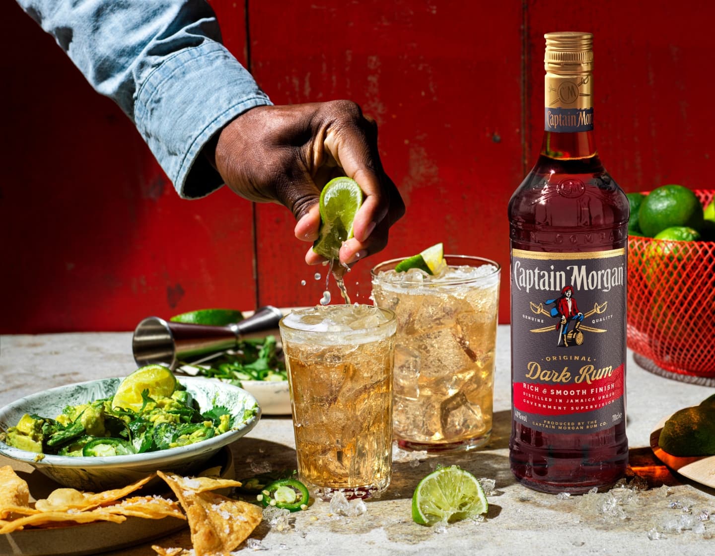Bottle of Captain Morgan Dark Rum on table surrounded by assorted food and drink against red background with a hand squeezing lime into one of the glasses