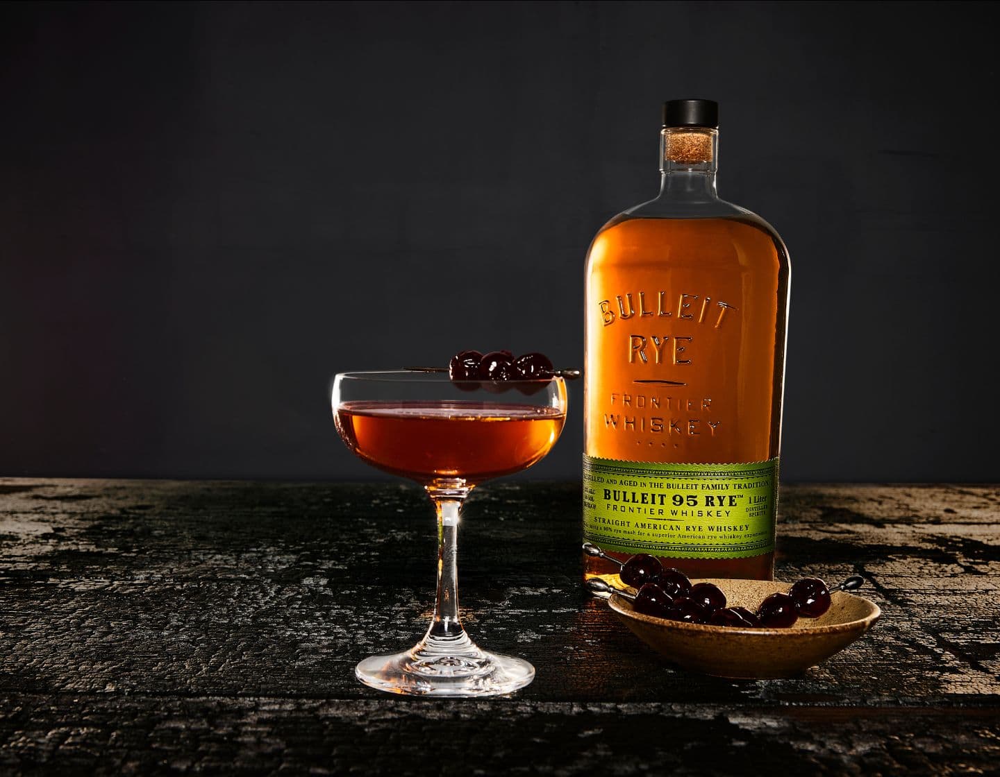 Bulleit_bourbon_bottle_beside_coupe_of_whiskey_cocktail_garnished_with_cherries