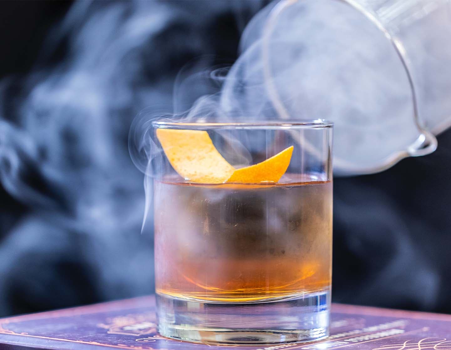 Um copo transparente cheio de líquido e uma casca de laranja rodeada de fumaça.