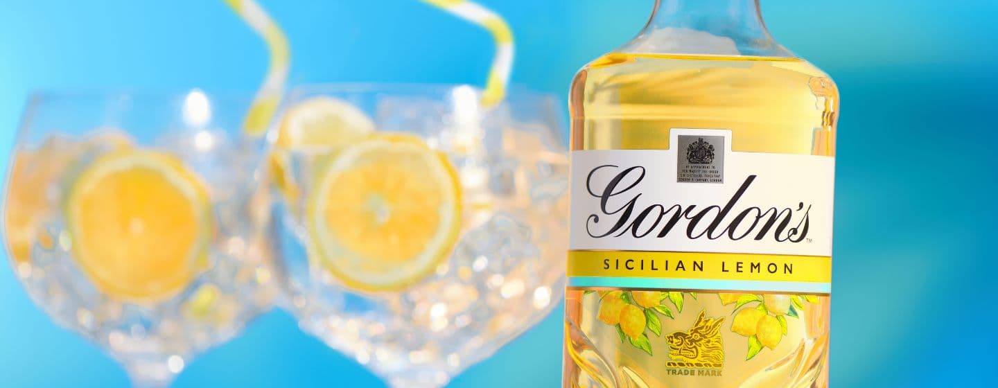  Una botella de ginebra Gordon’s Sicilian Lemon junto a dos copas de gin-tonic sobre un fondo azul 