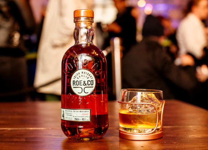 Botella de whisky Roe & Co sobre una mesa junto a un vaso de whisky con hielo 