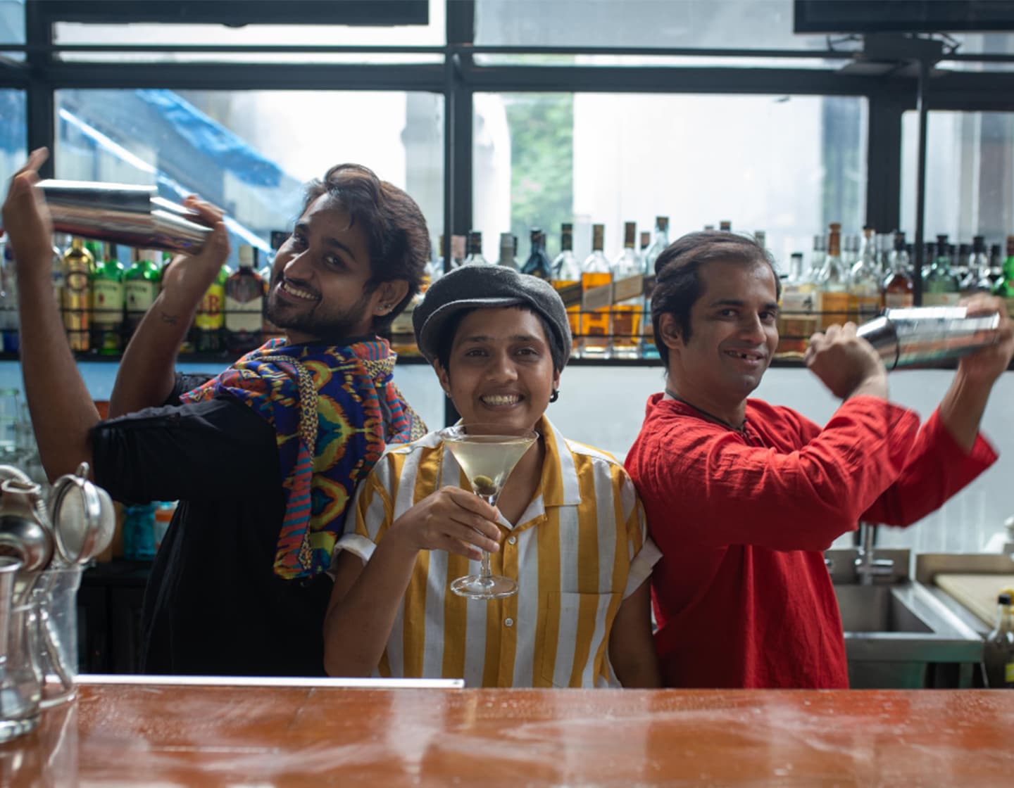 Three bartenders posed together, those on the outside holding cocktail shakers and smiling at the camera. The one in the middle lifting a cocktail glass and smiling at the camera. 