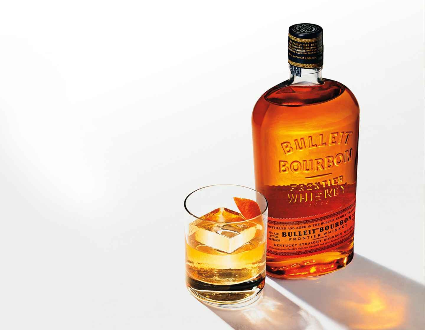 A bottle of Bulleit Bourbon on a white table next to a tumbler glass filled with Bulleit and a large cube of ice. Bright shadows are cast to the bottom right corner.