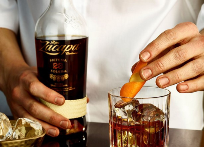 Mãos segurando uma garrafa de Zacapa e uma espiral de laranja sobre um copo de rum 