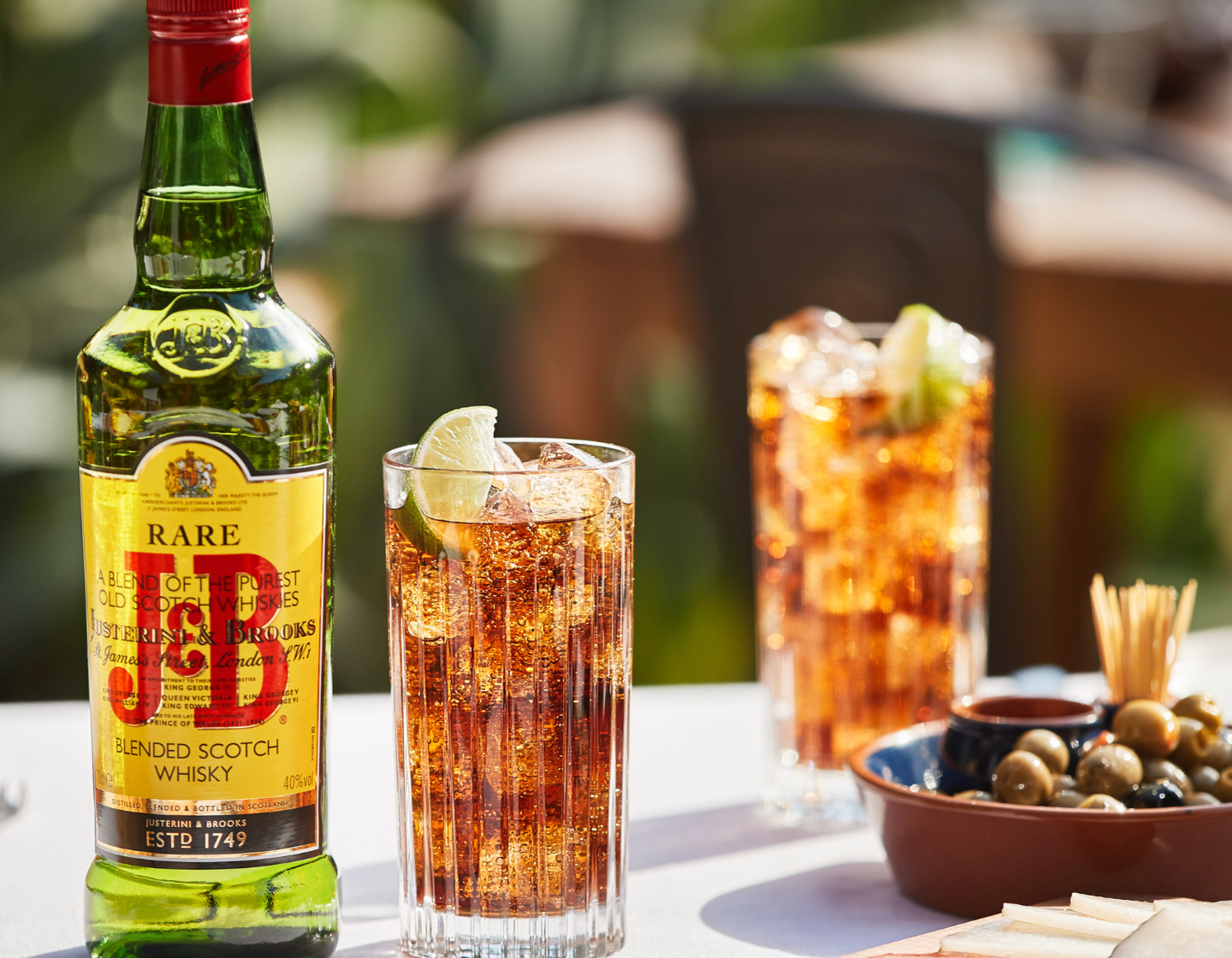 Botella de whisky J&B sobre una mesa de terraza con dos cócteles y un plato de aceitunas 
