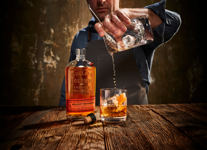 Man straining cocktail into a glass on table beside open bottle of Bulleit whiskey