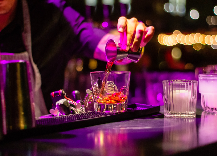 Eine Hand gießt an einer Bar eine Flüssigkeit aus einem Meßbecher in ein Getränk.