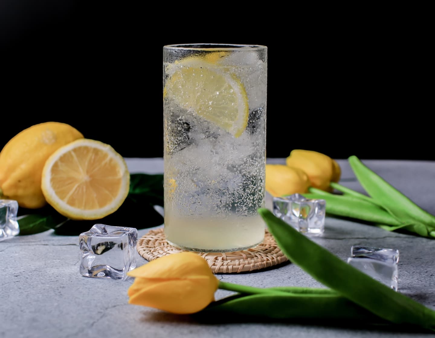 Cold drink in clear glass with sliced lemon and ice, placed on table with lemons ice and yellow flowers