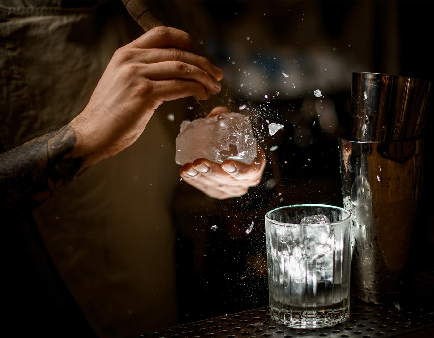 Imagem com iluminação fraca de duas mãos, uma segurando um grande cubo de gelo, a outra usando uma ferramenta de três pontas de lascar para criar um novo cubo de gelo com forma. 