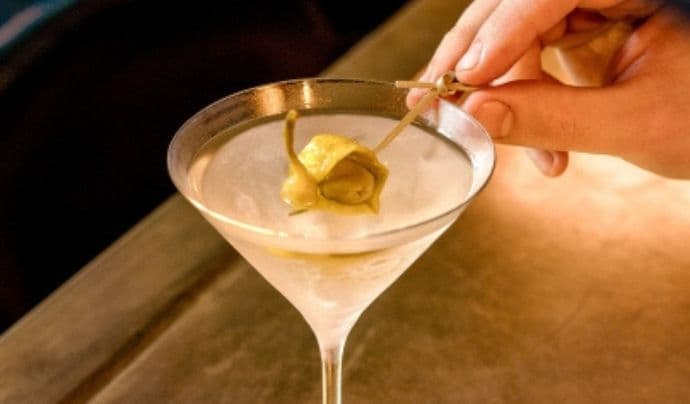 A martini drink with a bartender’s hand adding a green pickle garnish.