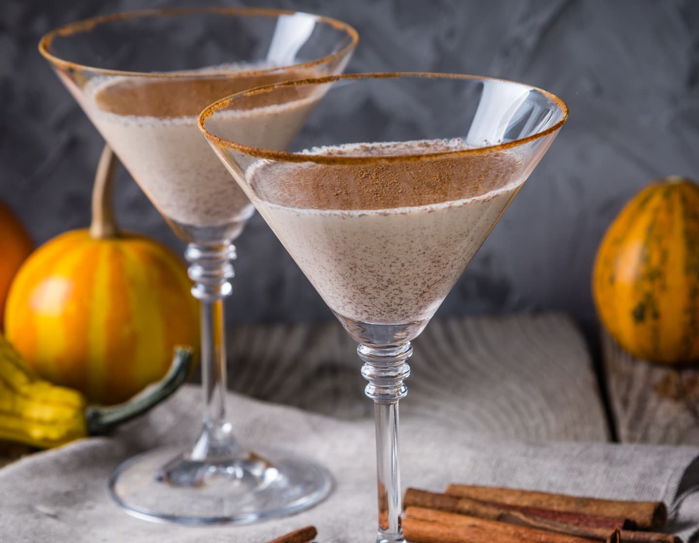 Two glasses of an autumn martini cocktail, with cinnamon sticks and small pumpkins surrounding them.
