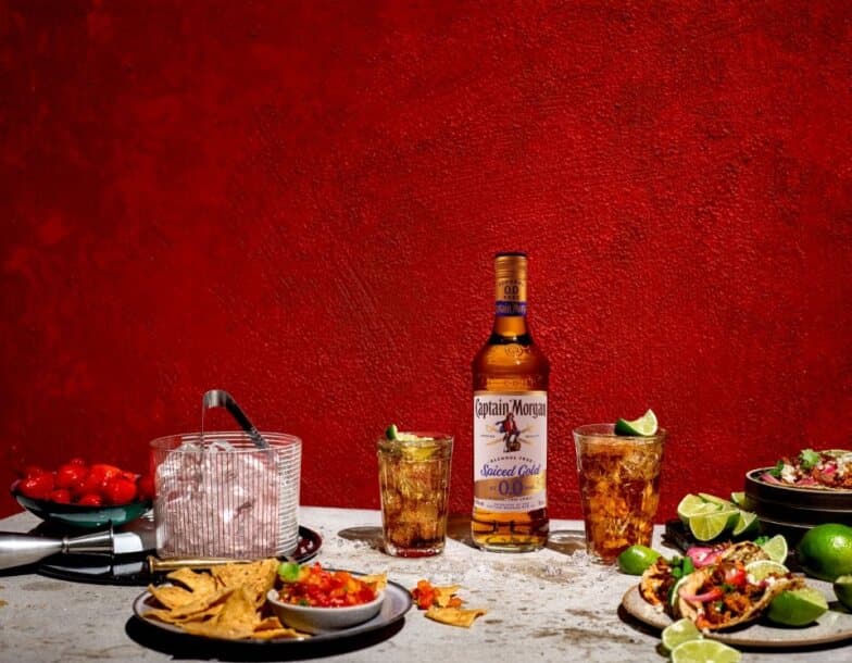 Botella de Captain Morgan Spiced Gold 0,0% sin alcohol sobre una mesa rodeada de alimentos y bebidas variadas sobre un fondo rojo.