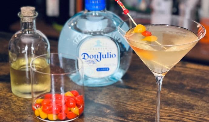 Un cóctel de martini teñido de naranja con guarnición de pepinillos rojos y naranjas y una botella de tequila Don Julio sobre una mesa de madera