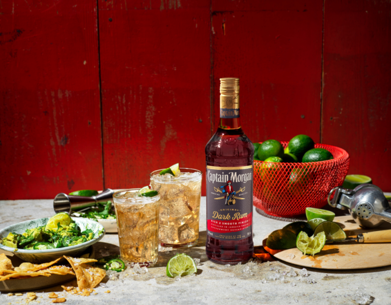 Botella de Captain Morgan Dark Rum sobre una mesa rodeada de platos y bebidas sobre fondo rojo. 