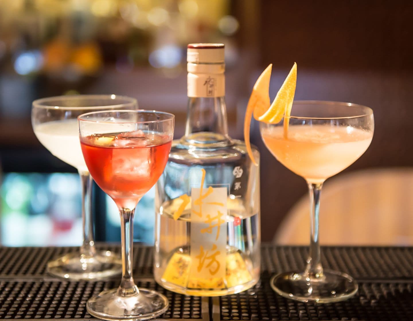 Eine Flasche Baijiu neben drei Cocktails auf einer Barplatte. 