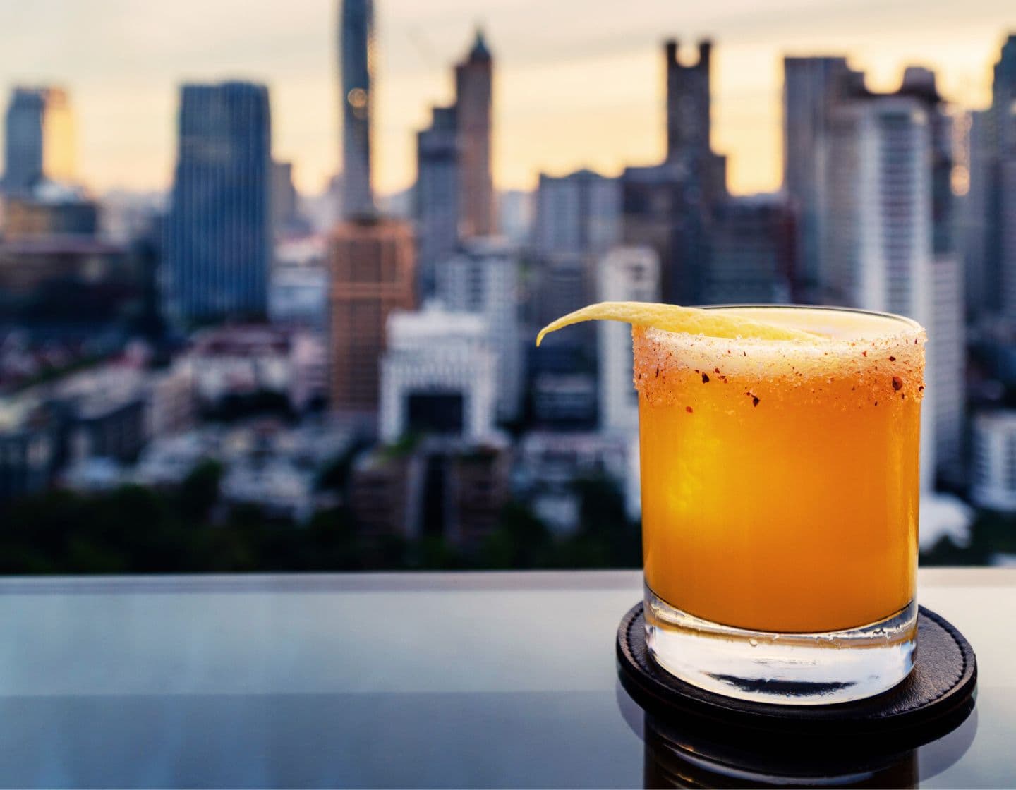 An orange cocktail garnished with lemon against a city skyline at dusk.