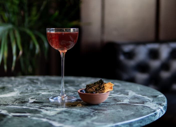 A red cocktail in a coupe glass with a small side dish of seaweed on a round, grey marble table with a plant in the background.