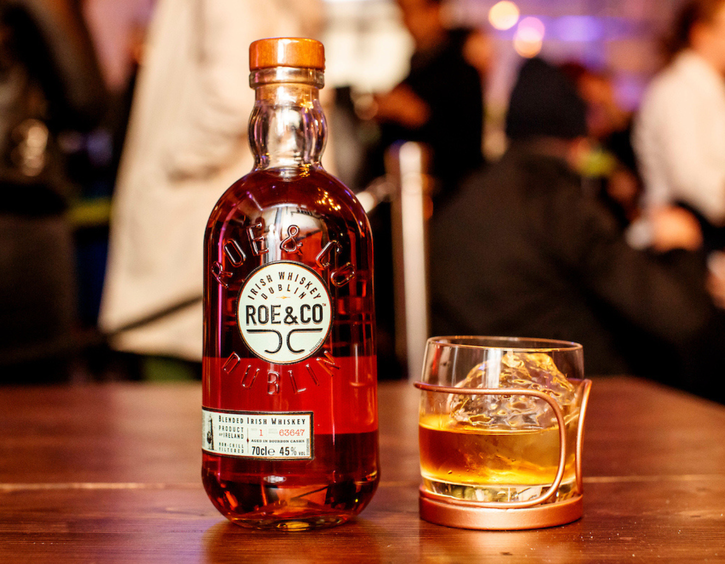 Botella de whisky Roe & Co sobre una mesa junto a un vaso de whisky con hielo 