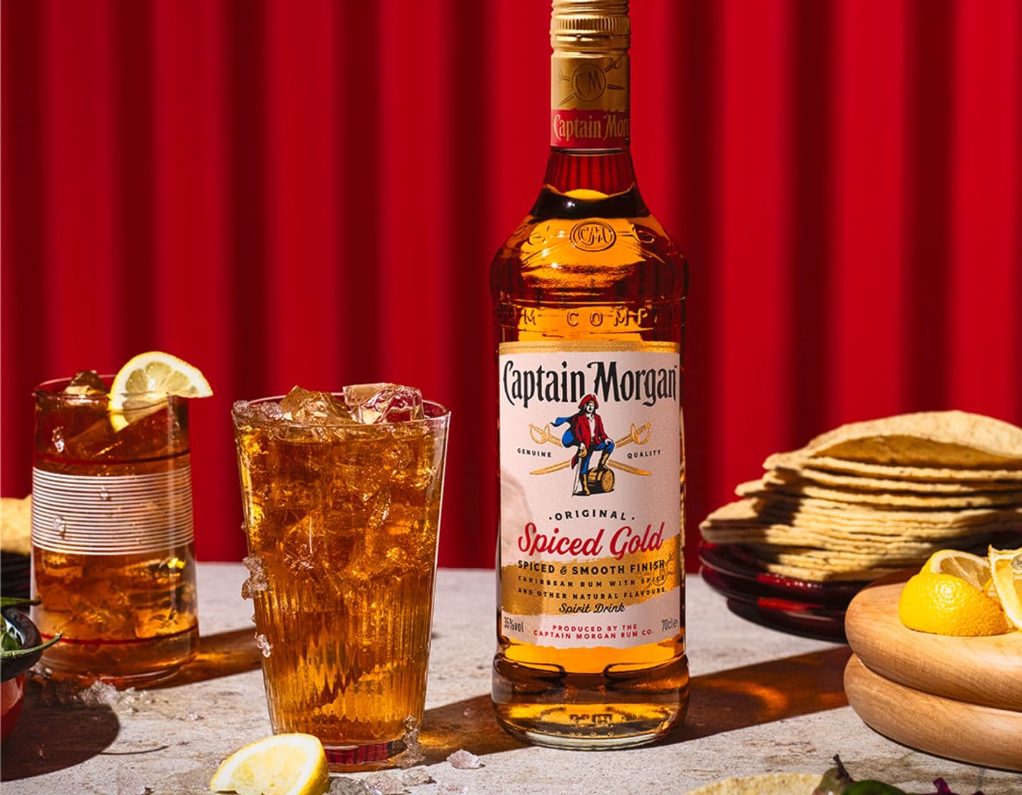 Flasche Captain Morgan Original Spiced Gold auf einem Tisch umgeben von verschiedenen Speisen und Getränken vor rotem Hintergrund