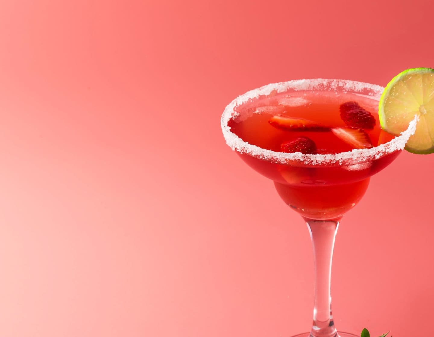 Red coloured Margarita cocktail with salt on the rim of a margarita glass and a lime wheel garnish. 