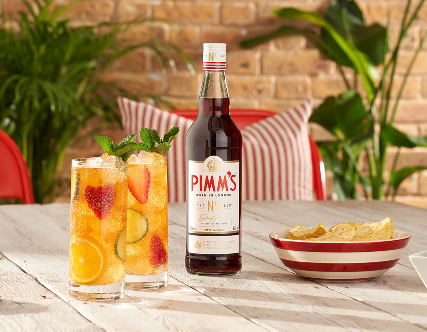 Botella de Pimm's sobre una mesa junto a dos vasos llenos de cócteles y un plato de papas fritas. 