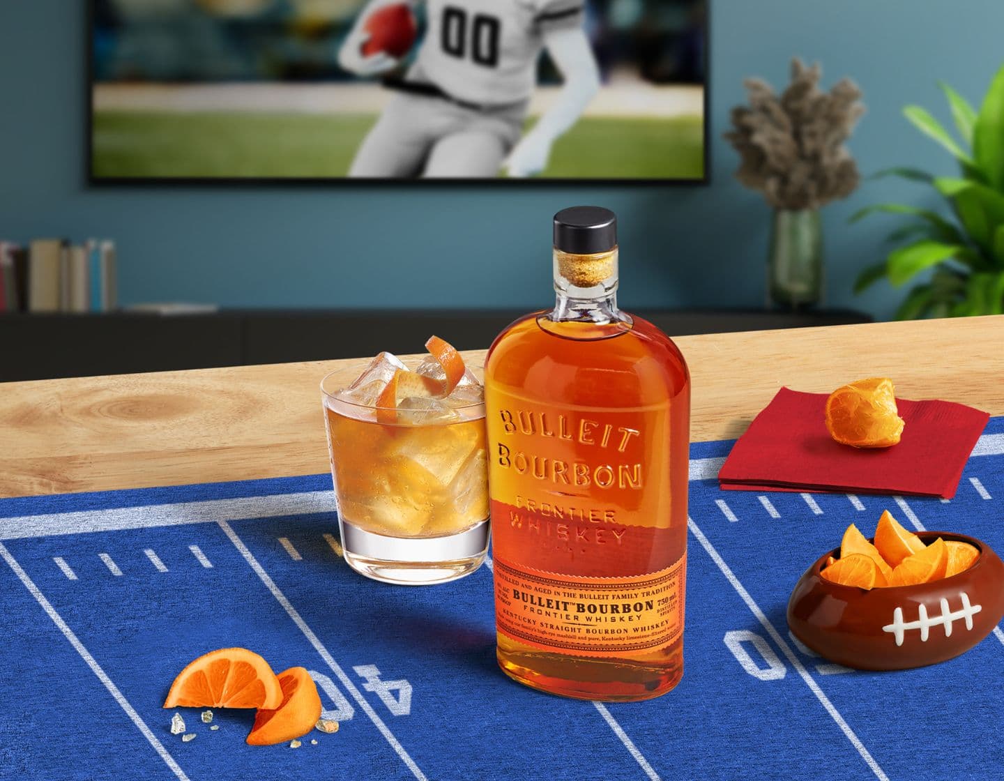 Bulleit_Bourbon_bottle_with_tumblr_glass_of_american_whiskey_and_oranges_on_table_with_sports_tv_in_background